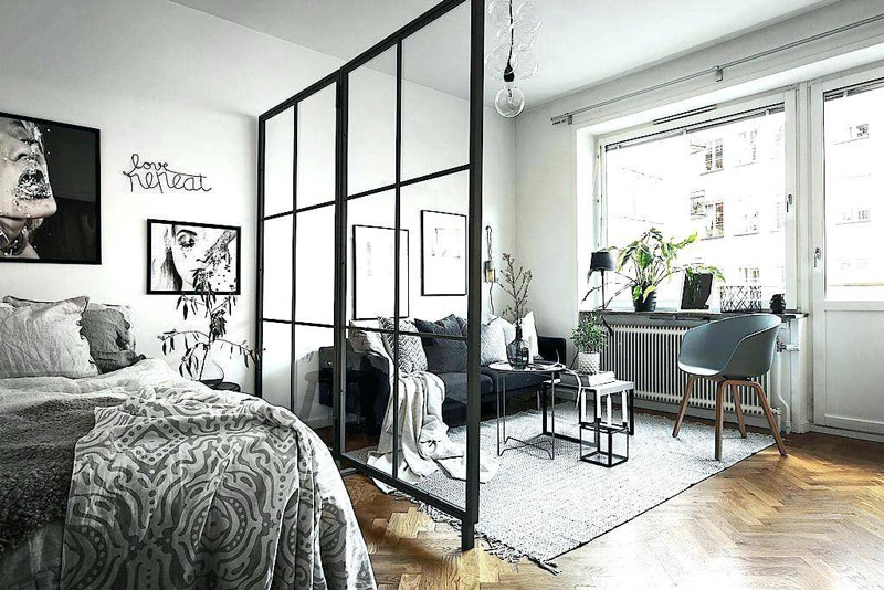 A well-lit bedroom with a bed on the left and a black-framed glass partition separating a sitting area with a couch and chairs. Large window with plants, minimalist decor, and framed art on the walls. For hire in Edinburgh: transport services like "man with a van" can assist in moving and setting up your space.