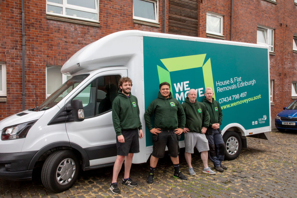 Four men in matching green hoodies standing in front of a parked moving truck on a cobblestone street beside a brick building in Edinburgh. The truck has "We Move" branding and contact details, offering reliable man with a van services for hire.