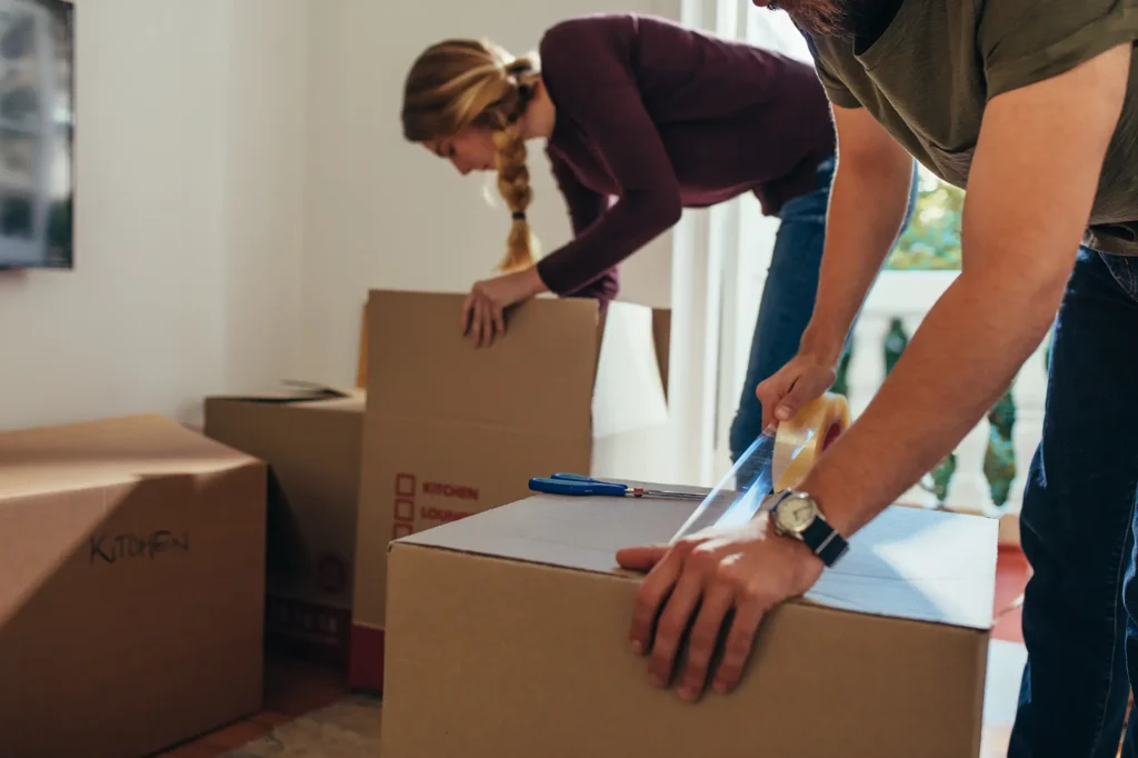 In a room bustling with activity, two individuals are packing boxes. One person is taping a box shut while the other is carefully arranging items in a box labeled "Kitchen." Outside, the man with a van for hire waits to transport their belongings across Edinburgh.