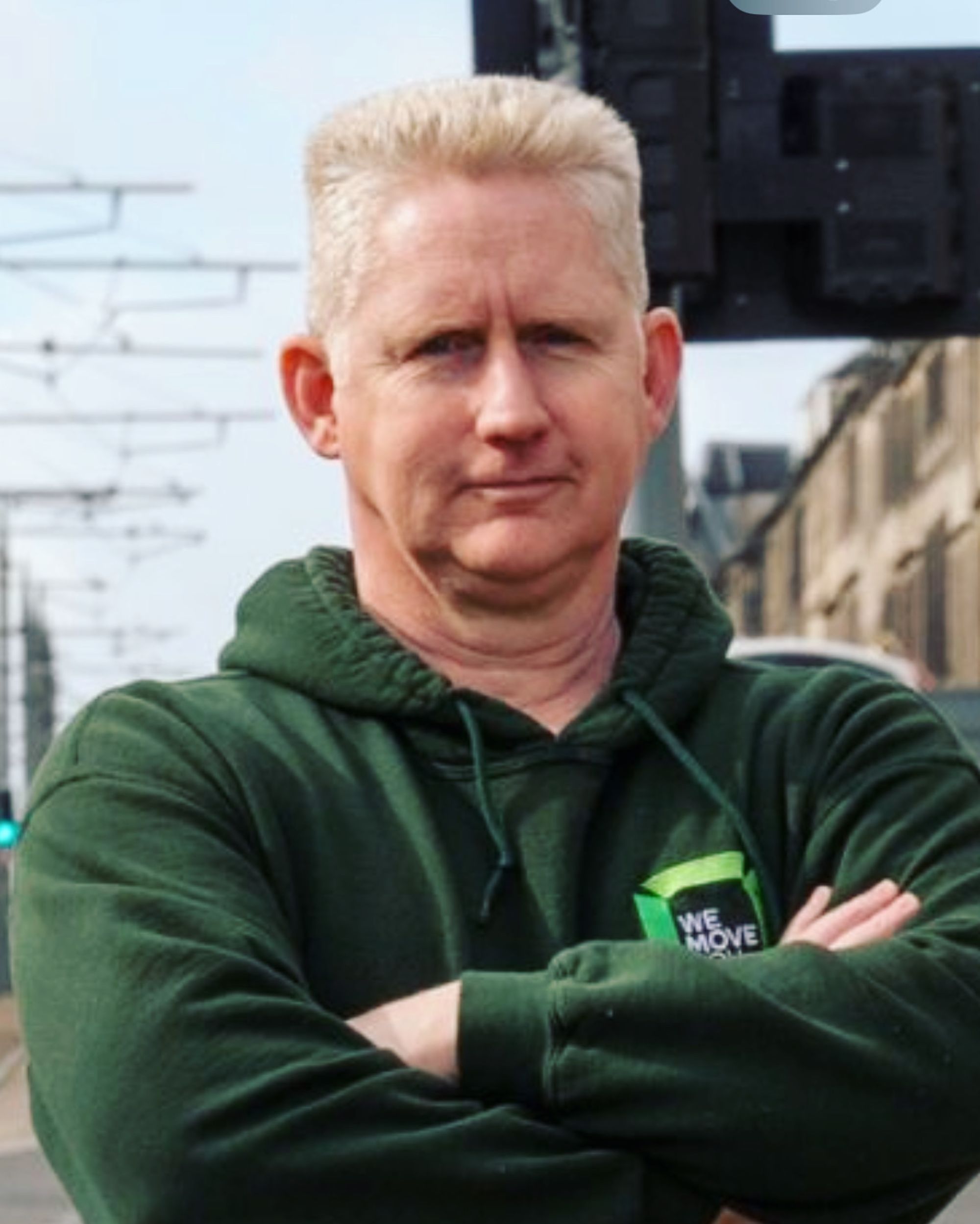 A person with short, light-colored hair wearing a green hoodie stands with arms crossed outdoors near buildings and railroad tracks in Edinburgh.