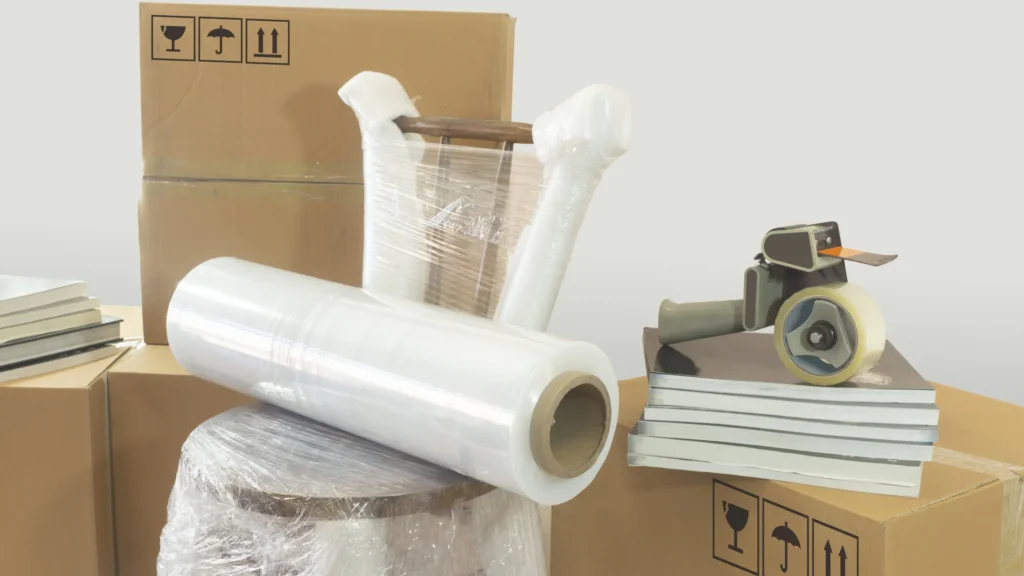 A chair and several boxes are wrapped in plastic. A packing tape dispenser and a roll of plastic wrap are placed on books, ready for the move. Additional cardboard boxes are in the background, awaiting the man with a van for hire to transport everything safely across Edinburgh.