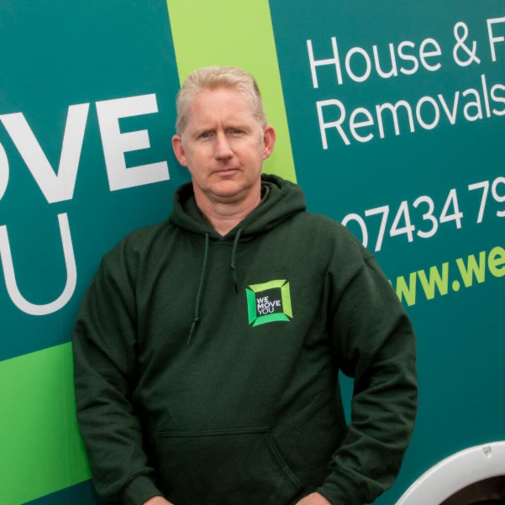 A man with grey hair wearing a green hoodie stands in front of a green and blue removals van for hire. The van displays the company's contact details and services in Edinburgh.