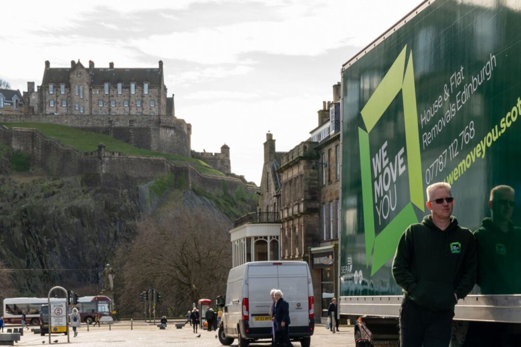 A person in a green hoodie stands next to a "We Move You" moving truck for hire. In the background, a historic stone building in Edinburgh sits atop a grassy hill. People and vehicles are on the street.