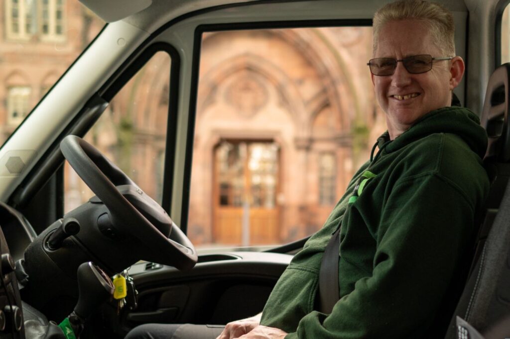 A smiling person wearing glasses and a green hoodie sits in the driver's seat of a vehicle, with a blurred building visible through the window. The man with a van is seemingly ready for hire in Edinburgh.