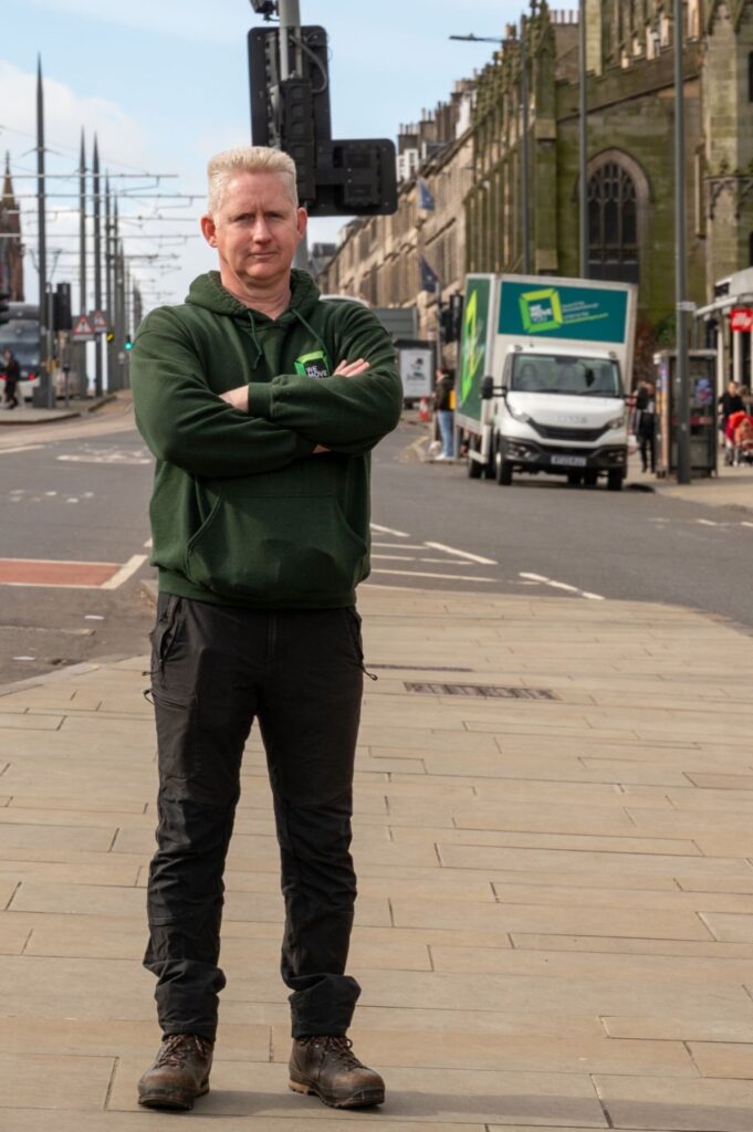 A man with short gray hair stands on an Edinburgh city street wearing a green hoodie and black pants, with arms crossed. A delivery truck labeled "Man with a Van for Hire" and historic buildings are visible in the background.