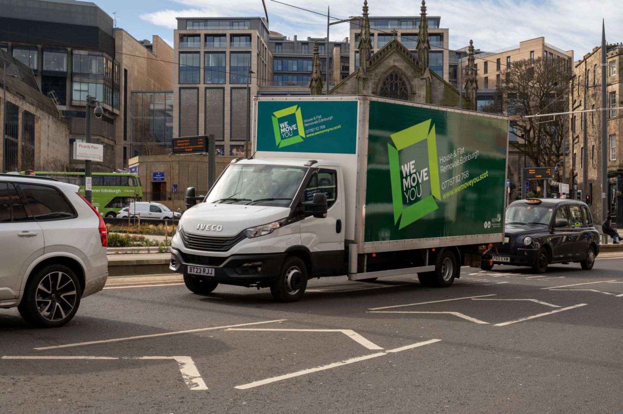 A delivery truck with "We Move You" branding, available for hire in Edinburgh, is driving down a city street, surrounded by other vehicles and modern buildings in the background.