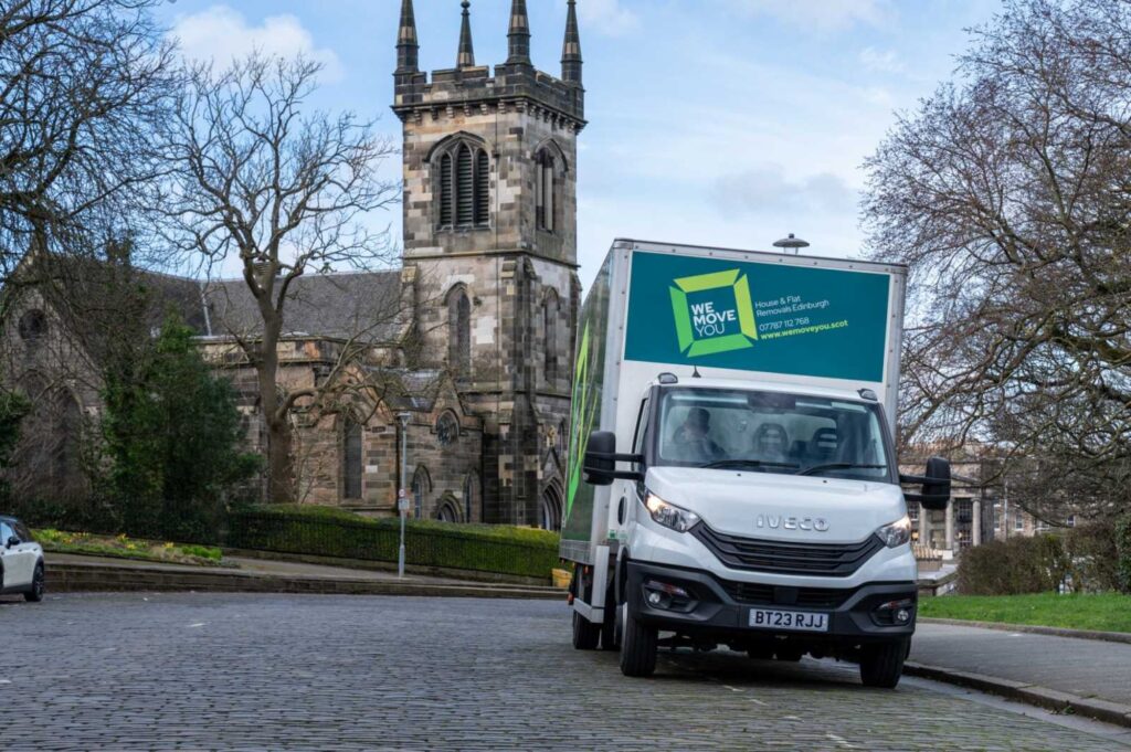 A delivery truck with a "We Move You" advertisement on its side, suggesting it's for hire, is parked on a cobblestone street near a stone building with a clock tower and spire in Edinburgh. Trees and blue sky are in the background.