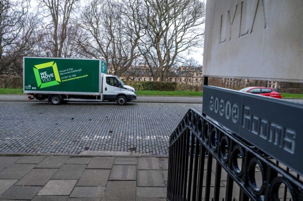 A green moving truck for hire is parked on a cobblestone street next to a building with "Rooms" written on a sign. Several trees and a few parked cars are visible in the background, capturing the charm of Edinburgh.