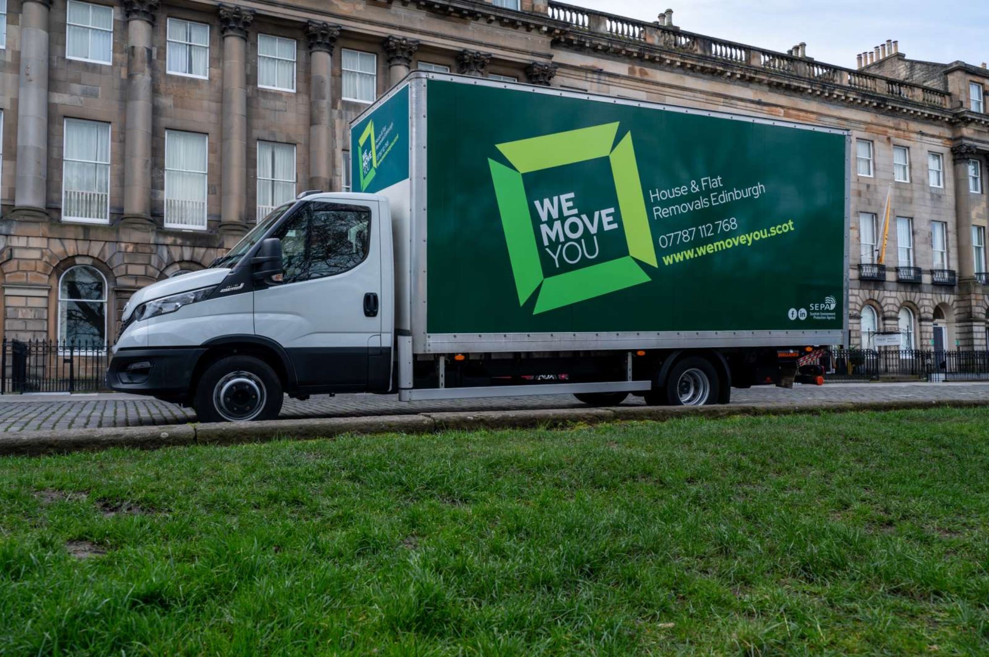 A green and white moving truck labeled "We Move You" is parked on a street in front of a row of buildings, offering reliable man with a van services for hire in Edinburgh.