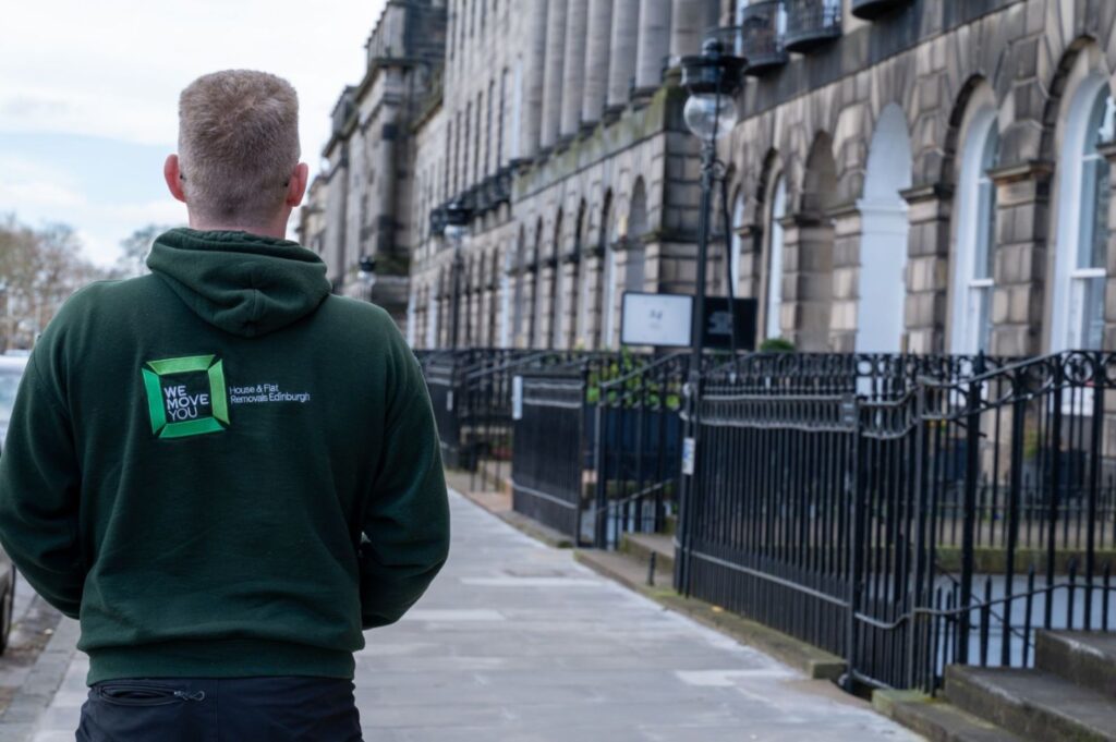 A person wearing a dark green hoodie with the text "We Move You" on the back walks on a sidewalk in front of historic buildings in Edinburgh. The scene gives the impression of someone ready for hire, perhaps a man with a van service.