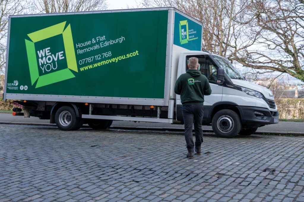 A person walks towards a green and white "We Move You" removals truck parked on a cobblestone street in Edinburgh. The man with a van offers house and flat removals, showcasing the service for hire along with a contact number and website.