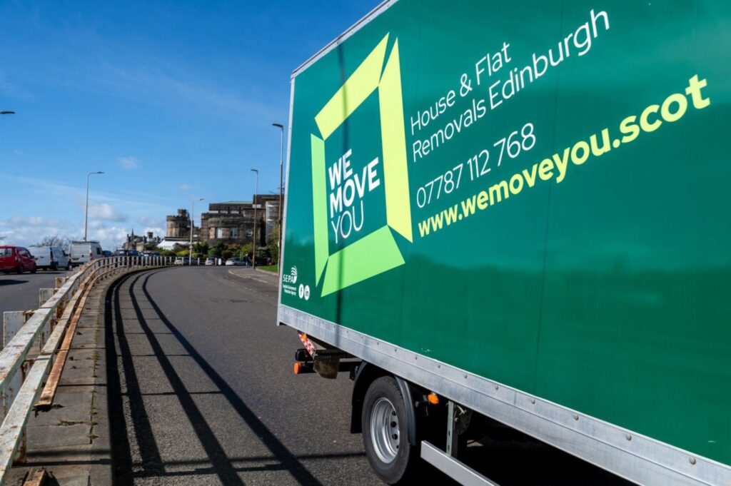 Green "We Move You" moving truck parked on a street with a fence on the left and buildings in the background. The truck advertises house and flat removals in Edinburgh, along with a contact number and website available for hire.