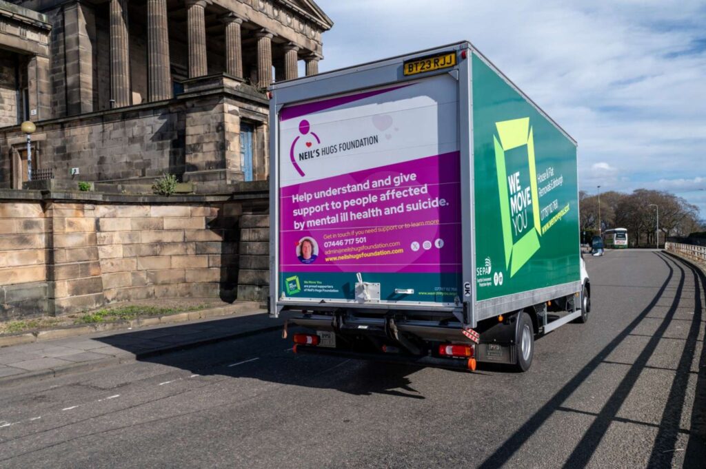 A delivery truck for hire, bearing a sign for Neil's Hugs Foundation—an organization supporting mental health and suicide prevention—is on a road next to an old stone building in Edinburgh.