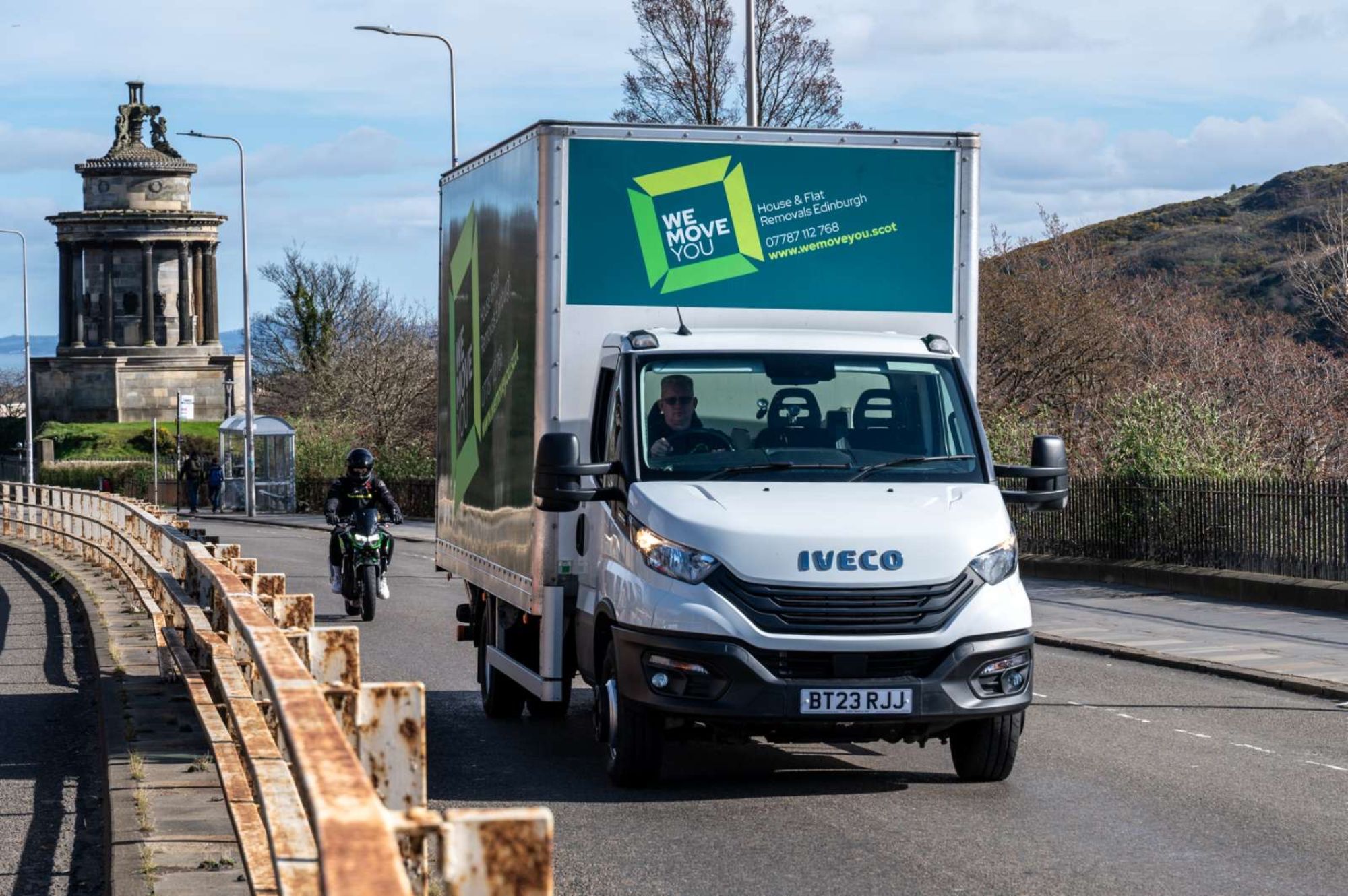 A white Iveco moving truck with the text "WE MOVE YOU" on the side is driving on a road in Edinburgh. A motorcycle follows behind. A classical monument is visible in the background, giving a hint of history to this "man with a van for hire" moment.