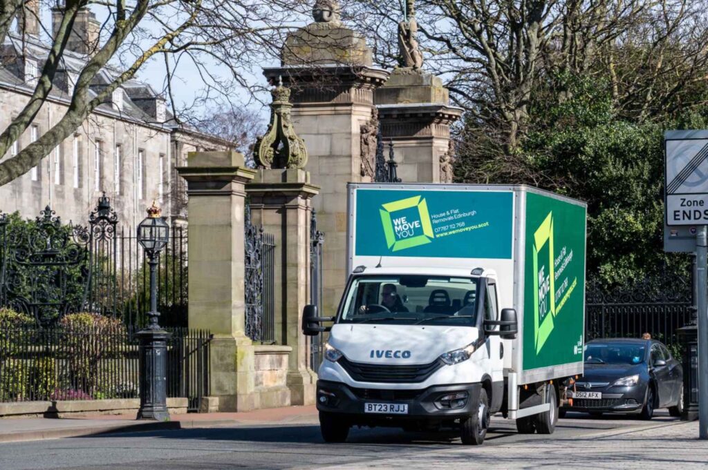 A man with a van for hire drives a green and white moving truck near stone columns and an ornate metal gate on a sunny day in Edinburgh.