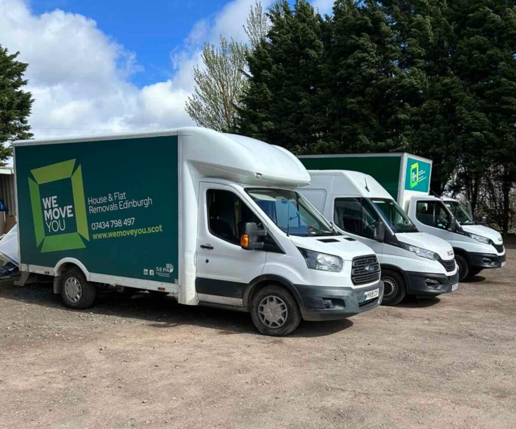 Three moving trucks labeled "We Move You" are parked outdoors on a dirt lot, with trees in the background. The trusted service is available for hire, ensuring your Edinburgh move is smooth and efficient.