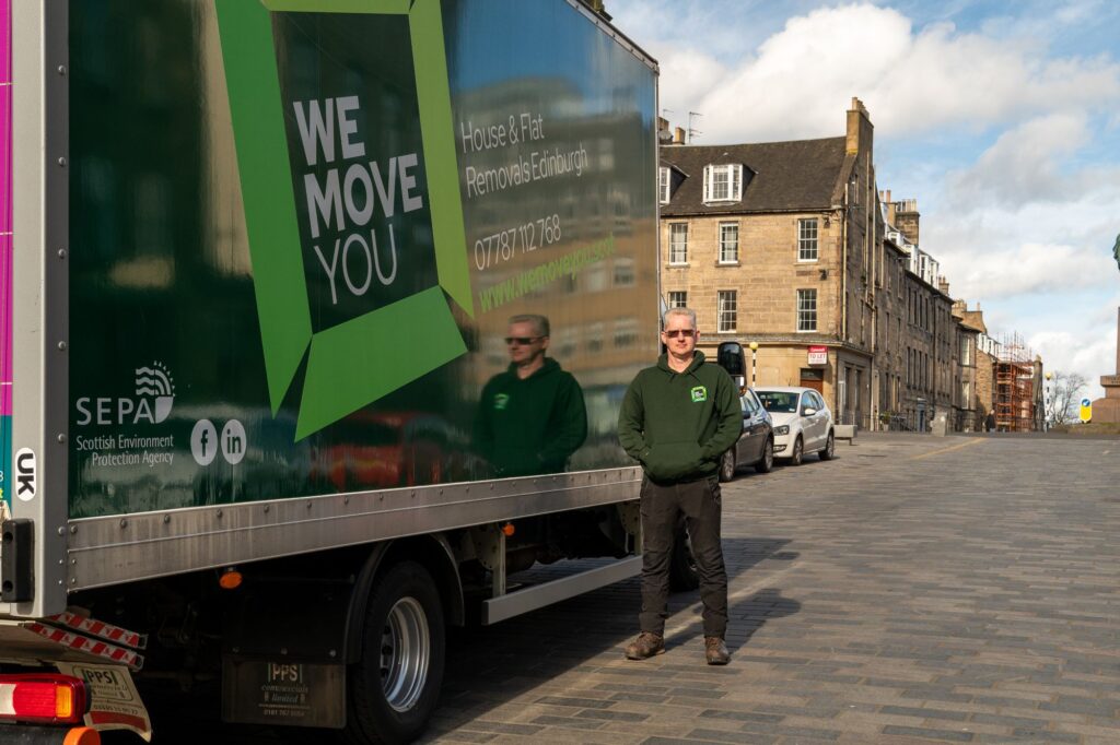 A man in a green hoodie stands beside a green moving truck labeled "WE MOVE YOU" on a cobblestone street in Edinburgh, with buildings in the background. The man with a van is ready for hire, blending seamlessly into the historic surroundings.